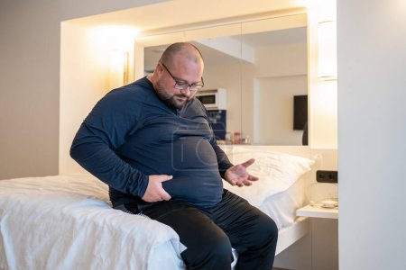 Photo for Huge man suffering from extra weight touching stomach sitting on bed in bedroom. Overweight bearded balded middle aged male with unhealthy body noticed problem. Weight control, overeating concept. - Royalty Free Image