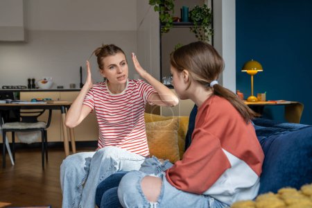 Young angry emotional mother screaming at troubled teen daughter, furious parent mom dealing with teenage behavior problems, yelling at upset adolescent girl while sitting together on sofa at home