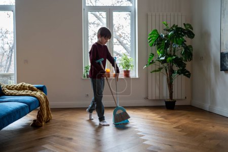 Photo for Teen boy doing chores cleaning floor in living room sweeping trash with broom to scoop. Teenager helping with household duties. Tidying up house involving children in family to maintain order at home. - Royalty Free Image