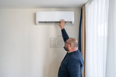 Téléchargez les photos : Homme vérifiant climatiseur de travail toucher l'air à la main en été temps chaud. Climatisation, système de séparation moderne dans l'appartement, appartement. Surchauffe mâle en surpoids se sent anormalement haute température de l'air. - en image libre de droit