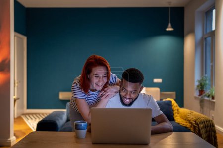 Photo for Interested smiling European woman looking to screen laptop with happy African American boyfriend. Pleased black man show funny video to wife. Happy couple reading good news in email, mortgage approval - Royalty Free Image