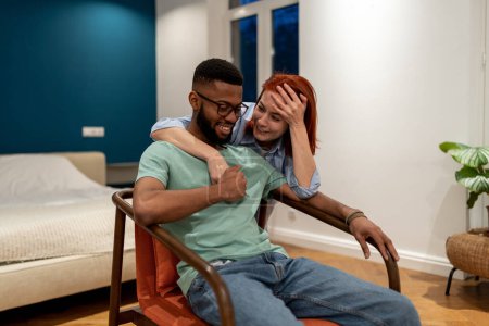 Photo for Overjoyed diverse couple speak each other relaxed family evening time at new rental home. Smiling African American man enjoy on armchair communication with hugging european laughing woman girlfriend - Royalty Free Image