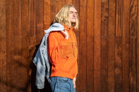 Photo for Relaxed European man in denim jacket over shoulder with close eyes stands near wooden wall in sun. Dreamy scandinavian blond guy freelancer enjoy sunny day take break resting outdoor near coworking - Royalty Free Image