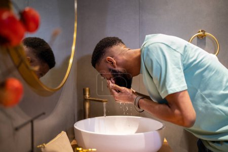 Photo for Skin care routine for men. Side view of African American millennial guy cleaning skin in morning, standing above bathroom sink and washing face with water, trying to wake up - Royalty Free Image