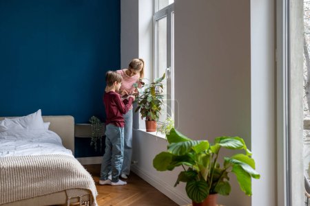 Photo for Mother teaching kid son how to care for plants and flowers, mom and interested boy standing near window at home examining leaves of houseplant. Parent explaining photosynthesis to child. Plant lovers. - Royalty Free Image