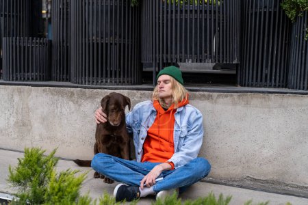 Foto de Molesto deprimido millennial chico sentado en la acera con el perro, tener problemas de vivienda. Un joven solitario en la calle de la ciudad pensando en problemas de vida. Desamparo y salud mental - Imagen libre de derechos