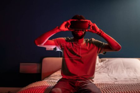 Téléchargez les photos : Homme noir utilisant casque de réalité virtuelle à la maison avec lumière au néon, assis sur le lit la nuit. Dépendance VR. afro-américain gars à l'aide de lunettes 3D, jouer à des jeux vidéo dans des lunettes, regarder étonné. - en image libre de droit