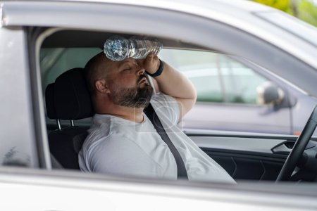 Photo for Overweight man drives car with broken air conditioner in hot summer weather. Weary male presses bottle of water to face to cool off suffering from heat, stuffiness. Exhausted tired overheated man. - Royalty Free Image