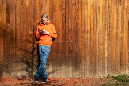 Photo for Full body portrait of blonde guy looks at smartphone screen stands near wooden high fence. Hipster with blonde long hair browsing internet, surfing online, reading message, chatting on smartphone. - Royalty Free Image