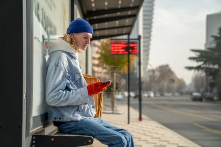 Téléchargez les photos : Étudiant du millénaire caucasien tenant smartphone assis sur un banc à la gare routière tôt le matin, attendant le transport, vérifier l'horaire de bus en ligne. Infrastructures de transports publics - en image libre de droit