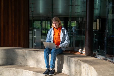Téléchargez les photos : Étudiant gars assis sur un banc de béton contre le bâtiment de l'Université, étudier en ligne à l'aide d'un ordinateur portable en plein air par temps ensoleillé. Homme millénaire pigiste avec ordinateur à l'extérieur. Freelance et urbanité - en image libre de droit