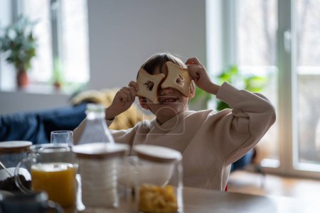 Photo for Son boy fooling playing with pieces of bread looking through holes at camera sitting at table on kitchen at home. Food eating, good mood cheerful playful child having fun during meal. Happy childhood. - Royalty Free Image