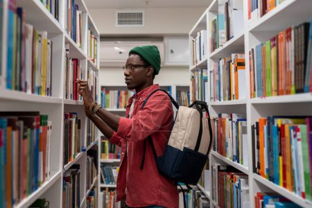Foto de Estudiante negro interesado en elegir un libro para leer en la librería. Enfocado chico afroamericano joven en busca de materiales para la investigación educativa en la biblioteca de la universidad. Literatura y educación - Imagen libre de derechos