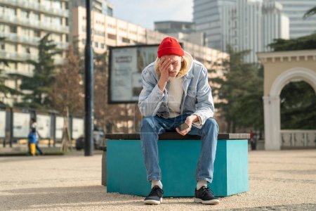 Photo for Depressed desperated guy hipster with phone sitting on bench in city having problems troubles. Frustrated confused man has bad news reads message on smartphone. Unfavorable life circumstances concept. - Royalty Free Image