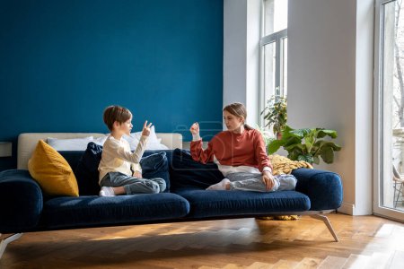 Photo for Children siblings sitting on sofa playing fun kids game Rock Paper Scissors to determine who has to clean room, brother and sister spending time together at home, family leisure concept - Royalty Free Image