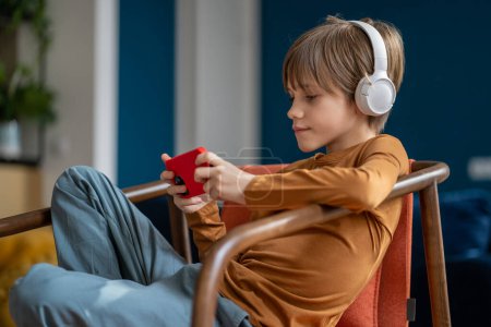 Photo for Children and screen time. Relaxed pre-teen boy child wearing headphones resting with smartphone at home, kid sitting on chair using mobile phone to listen to music or watch movies - Royalty Free Image