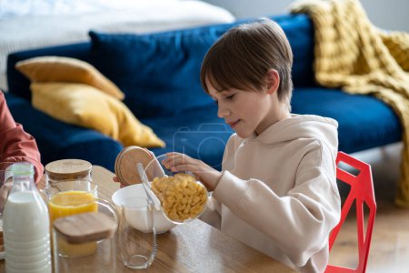 Photo for Teen boy preparing breakfast for himself pouring cornflakes in bowl sitting at table on kitchen. Healthy balanced meal for growing human body, independent schoolboy eating flakes at home. - Royalty Free Image
