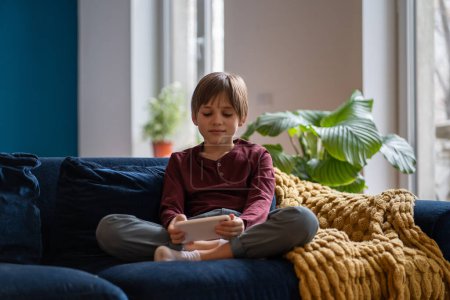 Foto de Niño pequeño jugando juegos móviles mientras descansa en casa, niño sentado en el sofá con teléfono inteligente en las manos. Un colegial mirando la pantalla del teléfono. Los niños y el concepto de adicción al juego - Imagen libre de derechos