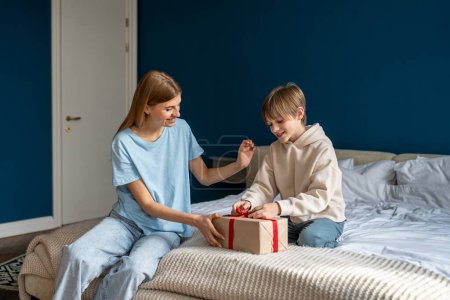 Photo for Cheerful kid son unwrapping gift box feeling excited getting present from caring mother, celebrating happy birthday. Loving family. Smiling mum congratulating child boy at home sits together on bed. - Royalty Free Image