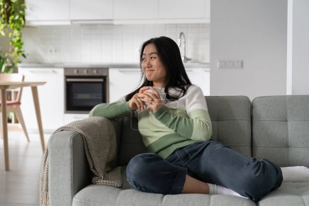 Pleased smiling Korean girl sits on sofa in home interior with cup of coffee in hands. Positive cheerful Asian woman enjoying day off, take break, looking away dreamily thinking about happy future
