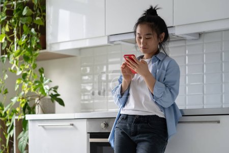 Mujer asiática optimista se para en la cocina mira mensajes de medios sociales de amigos y familiares con una leve sonrisa. Chica china complacido involucrado pasa tiempo libre de compras en la tienda en línea en el teléfono inteligente en casa