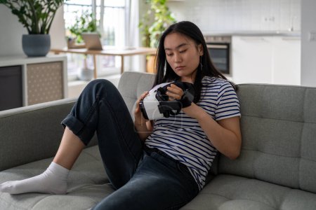 Téléchargez les photos : Focused femme chinoise assise sur le canapé considère lunettes de réalité virtuelle 3D pour l'immersion dans l'univers du jeu. Concentré asiatique jeune femme au repos à la maison en choisissant une nouvelle activité prêt à utiliser VR casque - en image libre de droit