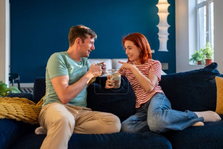 Foto de Feliz pareja de jóvenes casados relajados tomando café o té mientras descansan en el sofá en el apartamento moderno, la esposa y el marido hablando mientras se relajan en el sofá, se unen, disfrutan de un fin de semana acogedor en casa - Imagen libre de derechos