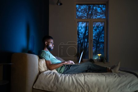 Photo for Focused African American programmer man in dark room lying on bed work on laptop. Workaholic freelancer black guy sitting comfortably with computer on laps working on graphic design for app at night - Royalty Free Image