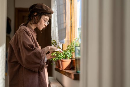 Hipster girl taking care of domestic houseplants, standing by window at home checking leaves and roots of indoors plants. Gardening and plant health care concept