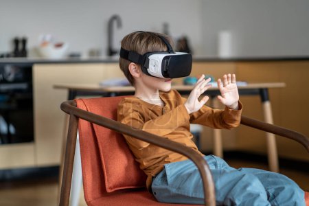 Téléchargez les photos : Enfant garçon portant un casque VR gesticulant avec les mains alors qu'il était assis sur une chaise à la maison. Les enfants interagissent avec le monde 3D, l'expérience de l'environnement virtuel. Enfants et sécurité en ligne - en image libre de droit