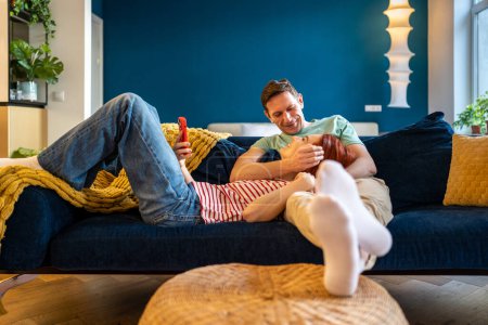 Photo for Relaxed happy woman lying with smartphone on husband knees while resting relaxing together on couch in living room. Married couple spending time with each other at home, embracing enjoy weekend - Royalty Free Image