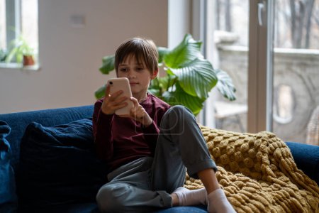 Relaxed little boy using mobile phone, looking at screen. Interested cute schoolboy sitting on couch at home with smartphone. Kid playing cellphone game, watch cartoons online. Child device addiction