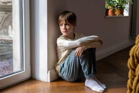 Photo for Kid psychological trauma. Lonely child boy sadly look to window hug knees sits on floor at home alone. Upset offended child thinking about family problems, bad relationship, failures with school peers - Royalty Free Image