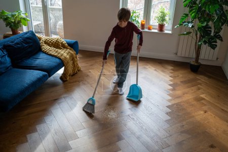 Photo for Independent child boy. Habit of cleaning house since childhood. Focused schoolboy sweeps floor from scattered debris with broom with brush and dustpan. Kid do house chores, domestic cleanup. - Royalty Free Image