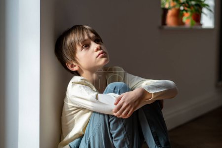 Photo for Lonely little boy sadly looking up hugging knees sits near window at home alone. Upset unhappy child waiting for parents, thinking about family problems, bad relationship. Kid psychological trauma - Royalty Free Image
