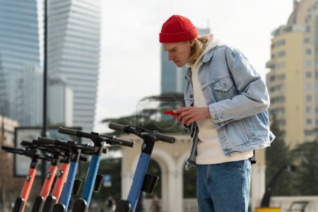 Photo for Interested Scandinavian hipster guy explores scooter rental smartphone app for eco-friendly urban mobility. Focused concentrated young european man scans qr code of individual transport in city center - Royalty Free Image