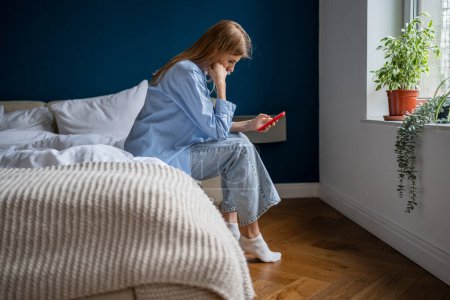 Photo for Depressed female holds cellphone pondering bad offer worried think about problem difficult solution trouble. Frustrated woman sits on bed feels upset reads bad news in message on smartphone at home. - Royalty Free Image