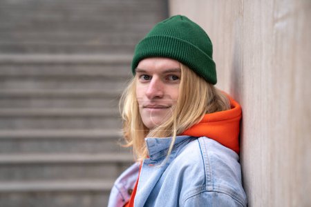 Téléchargez les photos : Portrait urbain d'un homme hipster millénaire heureux aux longs cheveux blonds souriant à la caméra, appuyé sur un mur de béton tout en se tenant debout sur des escaliers dans un passage souterrain. Vie urbaine et bonheur - en image libre de droit