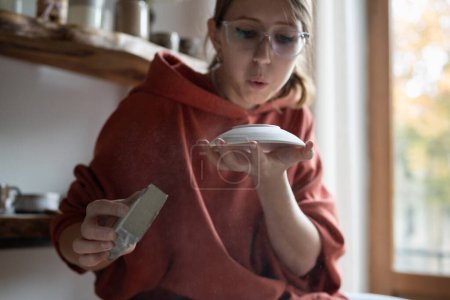 Photo for Immersive in process girl blowing on porcelain plate to remove excess dust sitting in craft workshop. European woman in glasses employee of craft workshop holding polishing sponge sits near window - Royalty Free Image