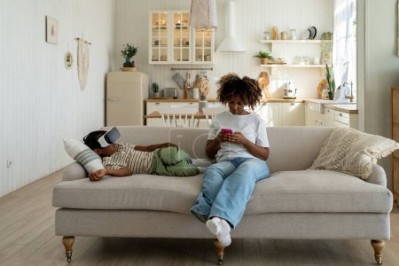 Foto de Madre afroamericana ocupada enfocada sentada en el sofá mirando el teléfono inteligente de la pantalla mientras su hijo pequeño usa gafas 3D de realidad virtual. Relajado colegial viendo película con gafas VR cerca de mamá en casa - Imagen libre de derechos