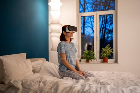 Foto de Chica se sienta en la cama en gafas de realidad virtual. Mujer juega videojuegos a través de simulador de casco especial. Turista en hotel con dispositivo especial para VR explora la ciudad. Futuro ahora - Imagen libre de derechos