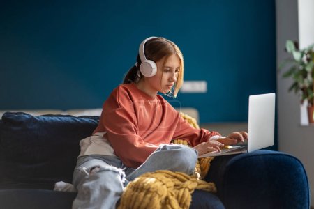Téléchargez les photos : Une adolescente concentrée portant un casque tapant du texte sur un ordinateur portable détendue assise sur un canapé dans le salon. Pensive adolescent écolier étudiant en ligne, enseignement à distance, en utilisant l'ordinateur à la maison. - en image libre de droit