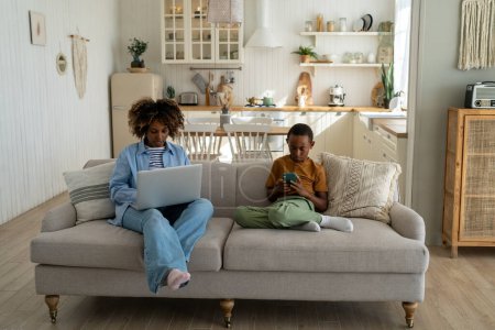Téléchargez les photos : Technologie et famille. Mère afro-américaine et son fils assis sur le canapé et regardant dans le téléphone et l'ordinateur portable. Dépendance au jeu chez les enfants. Maman occupée et enfant au téléphone. Parent au travail, garçon avec mobile - en image libre de droit