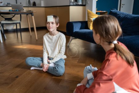 Photo for Smiling teen sister and child brother play together who am i guess sticker game sit on floor at home in living room. Girl has sticker with frog word on forehead. Family funny time, siblings friendship - Royalty Free Image