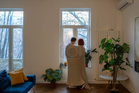 Photo for Family couple wife husband hugging looking at window standing in living room at home covering with knitted plaid, rear view. Affectionate man and woman in love cuddling together in cozy interior. - Royalty Free Image