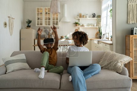 Foto de Afroamericana familia mamá y el hijo se sientan en el sofá con dispositivos electrónicos. Madre-padre se sienta con el ordenador portátil, hijo en gafas de realidad virtual. Negro chico olas manos en el casco de juego. Familia y tecnología - Imagen libre de derechos