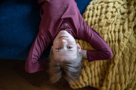 Téléchargez les photos : Portrait adolescent couché sur le canapé à l'envers avec les mains derrière la tête et la caméra de regard. Souriez sur le visage d'un enfant. Son fils se repose avant l'école. Journée internationale de l'enfant - en image libre de droit