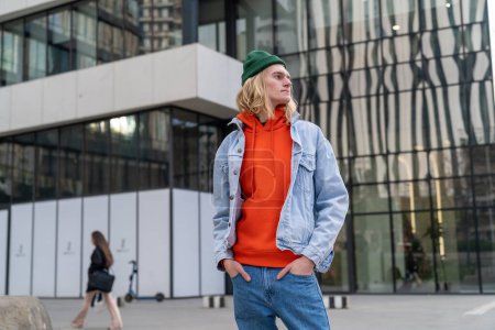Photo for Portrait oeuropean young hipster man, hands in jeans pockets, looking sideways and standing next modern business center in big city. Stylish blond male in denim near shopping malll. Urban lifestyle - Royalty Free Image