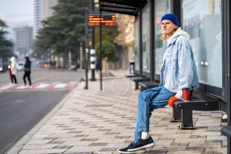 Photo for Young blond guy student waiting transport very long time in morning.Man on bus stop with coffee away. Blonde hair millennial man on bus stop in city. Modern urban transport for everyone - Royalty Free Image