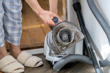 Female opening dust filter out of vacuum cleaner at home. Hoover container full of dirt, fur, pet hair after cleaning at home. Dust in apartment is source of allergies. Household chores concept
